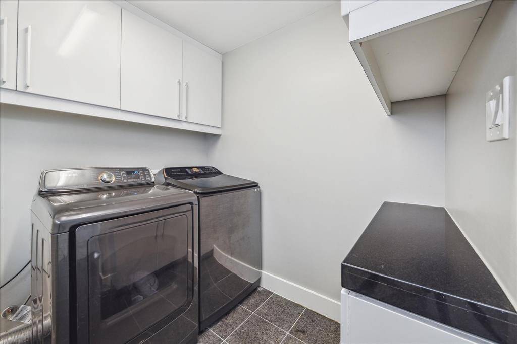 Modern laundry room featuring a sleek washer and dryer set with stainless steel finishes. It has ample storage with upper cabinets and a countertop for folding or organizing. The space is bright with neutral walls and tiled flooring.