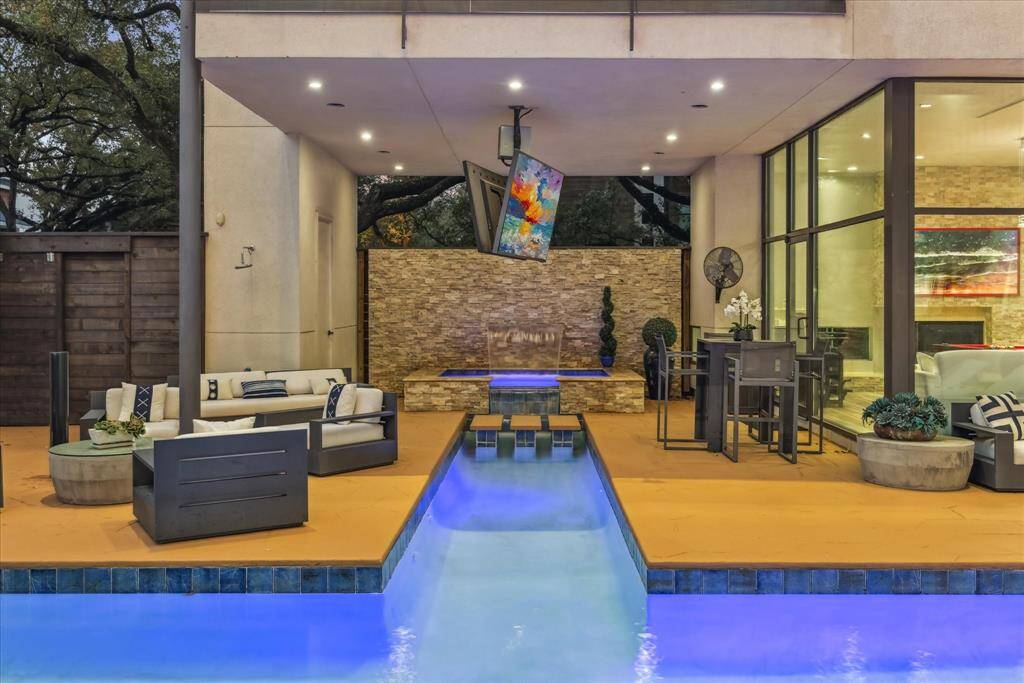 This outdoor space features a modern patio with a cozy seating area, a sleek pool with vibrant lighting, and a covered section with a mounted TV and dining set. The design offers a seamless indoor-outdoor flow, perfect for relaxation and entertaining.