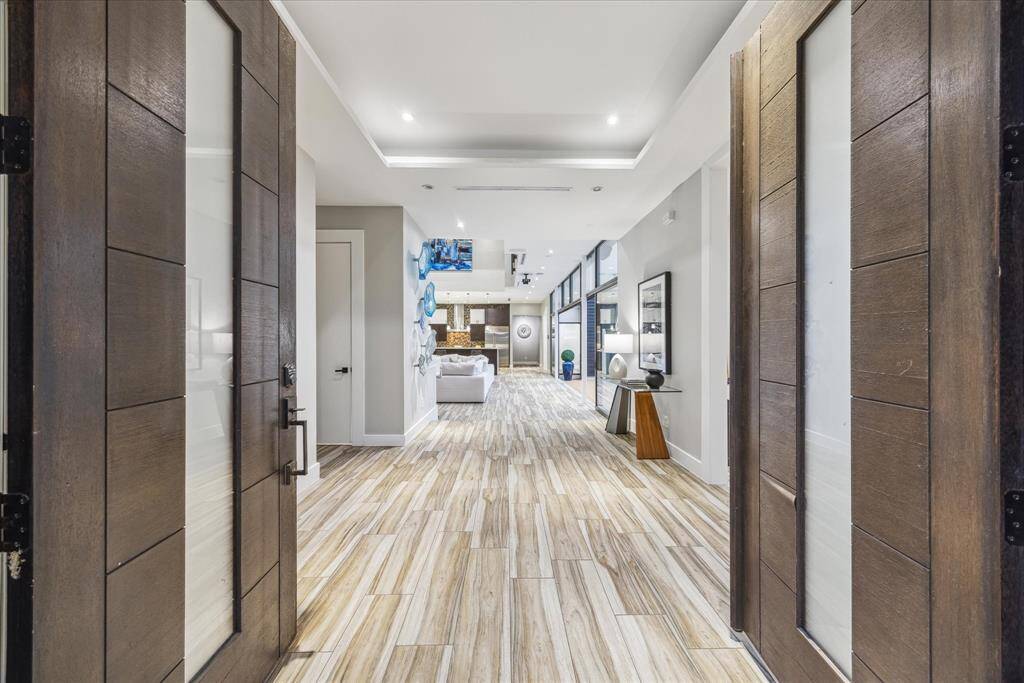 Welcome in! A modern, inviting entryway with wide wooden double doors leading into a bright hallway. The space features sleek, light wood flooring, and recessed lighting creating a welcoming atmosphere.