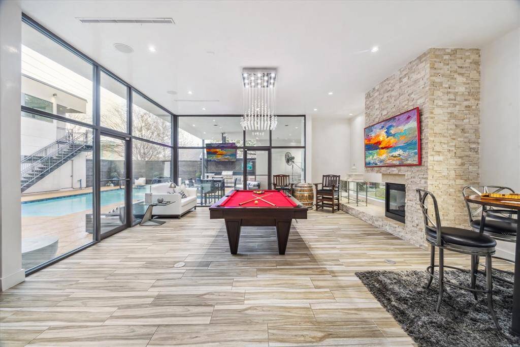 This modern living space features a large, open area with elegant chandelier, and sleek flooring. It has expansive windows offering views of a pool, a cozy fireplace with a stone accent wall, creating a stylish and inviting atmosphere.