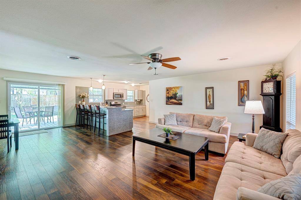 This open-concept living area features hardwood flooring and ceiling fan. The modern kitchen offers an island with bar seating and stainless steel appliances. Sliding glass doors lead to a patio, enhancing natural light and outdoor access.