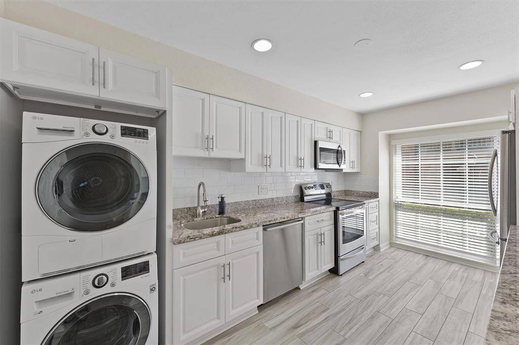 This modern kitchen features white cabinetry, granite countertops, and stainless steel appliances. It includes a stacked washer and dryer, a spacious sink, and a large window providing ample natural light.