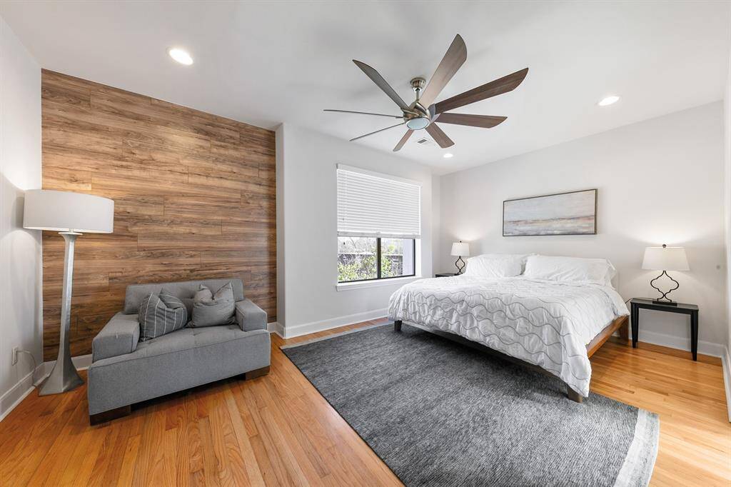 Located down a long, private hallway off the upstairs den, the primary bedroom suite immerses the homeowners in peace and tranquility at the end of the day, offering plenty of space for a library or private lounge.  Architecturally intriguing, it features a wood accent wall, red oak hardwood flooring, a striking contemporary ceiling fan, and recessed lighting.