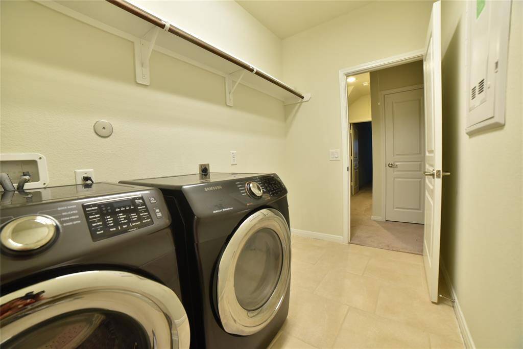 This laundry room features modern front-loading washer and dryer units, tiled flooring, a shelf for storage, and neutral walls. It opens to a hallway, providing easy access to other parts of the home.