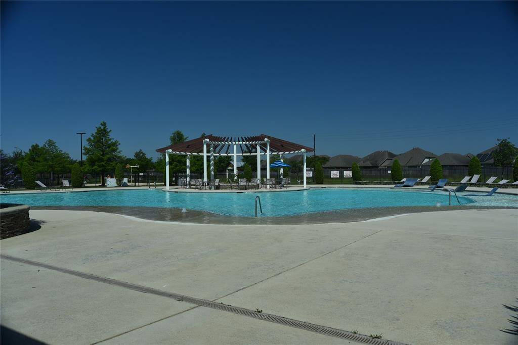 A spacious community swimming pool with lounge chairs and a covered seating area, set against a backdrop of greenery and neighboring houses, perfect for relaxation and gatherings.