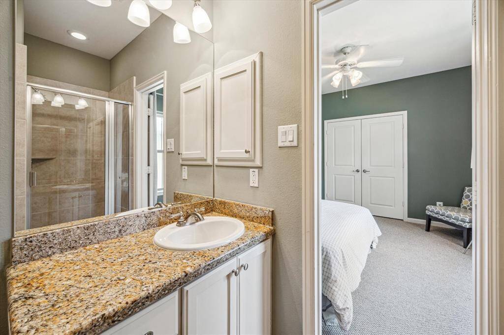 Granite countertops with warm, golden tones add texture and elegance to the en suite bath. A large framed mirror and soft overhead lighting brighten the space, while a walk-in shower with swing glass door offers a clean, modern finish. Custom cabinetry provides functional storage without sacrificing style.