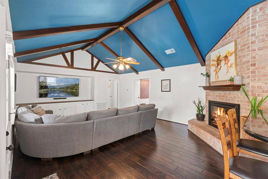 This inviting living room features a high vaulted ceiling with exposed beams, a striking blue color, and includes a cozy brick fireplace. The space is accented with dark hardwood floors and an overhead ceiling fan, creating a warm and stylish atmosphere.