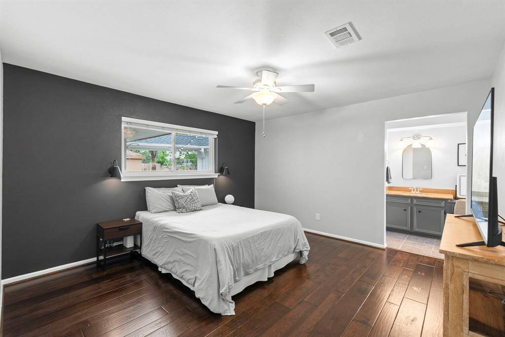 The bright and spacious primary bedroom features a dark accent wall, hardwood floors, and a ceiling fan. It includes a large window for natural light and an en-suite bathroom with modern fixtures.
