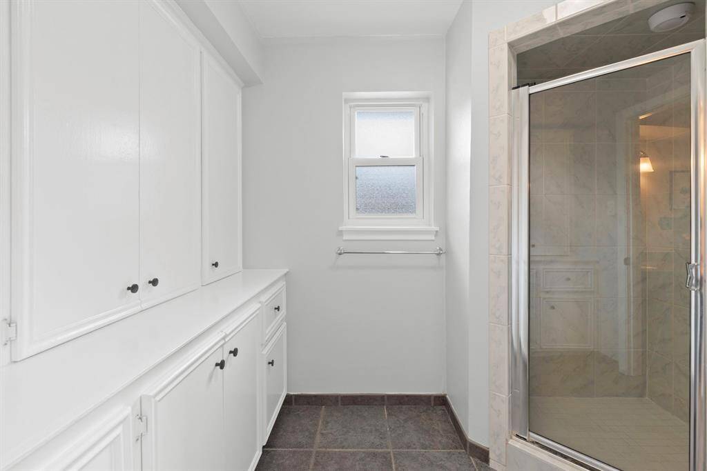 The primary bathroom also features ample white cabinetry for storage, a window for natural light, a glass-enclosed shower, and tiled flooring.