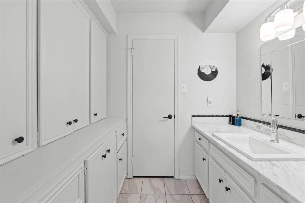 This bathroom features a clean, white design with ample cabinetry for storage, a large mirror, and modern lighting. The countertop is sleek with a single sink, offering a functional and stylish space.