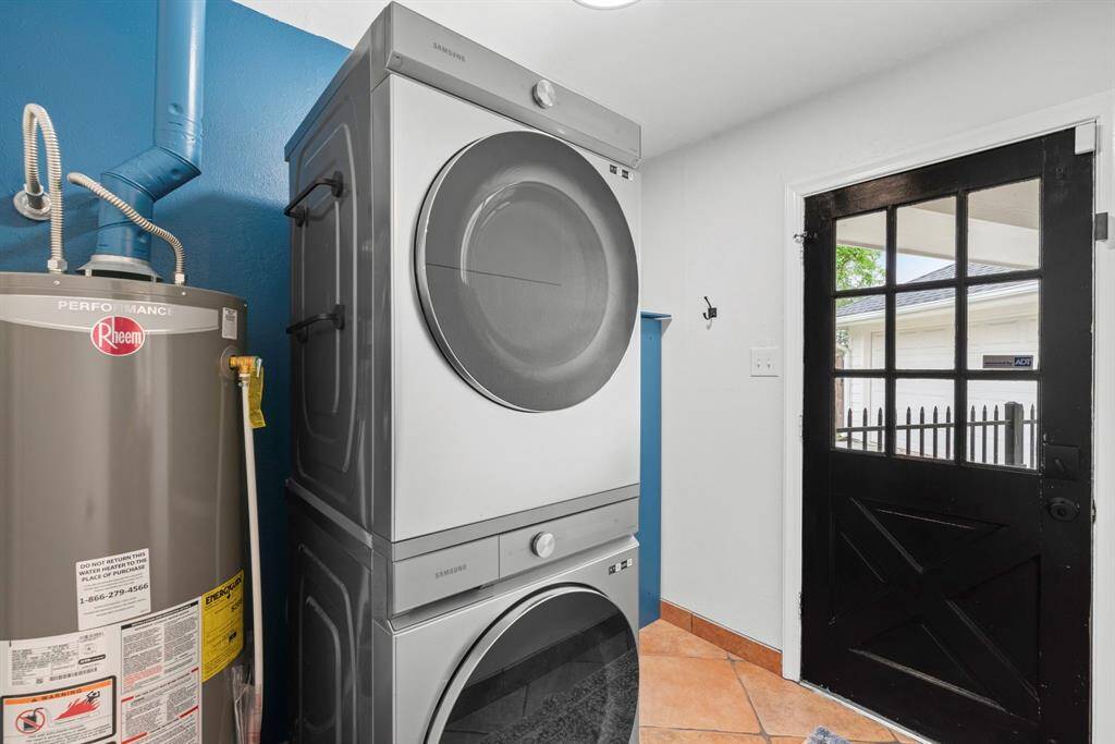 This photo shows a compact laundry room with a stacked washer and dryer next to the water heater. The space features a bright blue accent wall, terracotta tile flooring, and a black door with a window, leading outside. To the left of the water heater is the 1/2 bathroom.