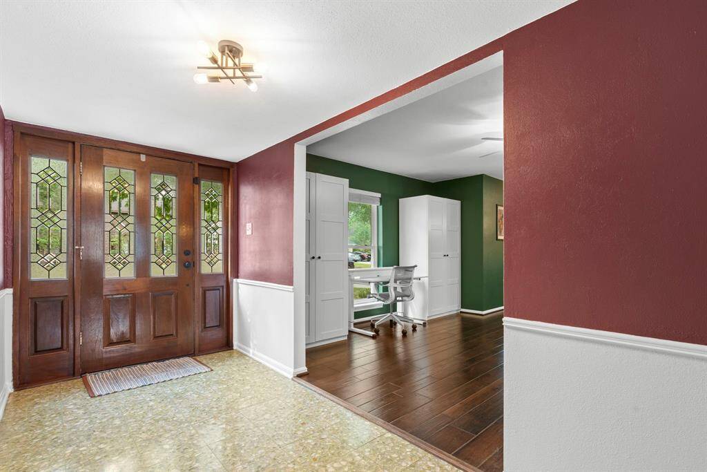 This photo showcases a welcoming entryway with a stylish wood and glass door, leading to a cozy formal living room featuring rich green walls and warm wooden flooring.