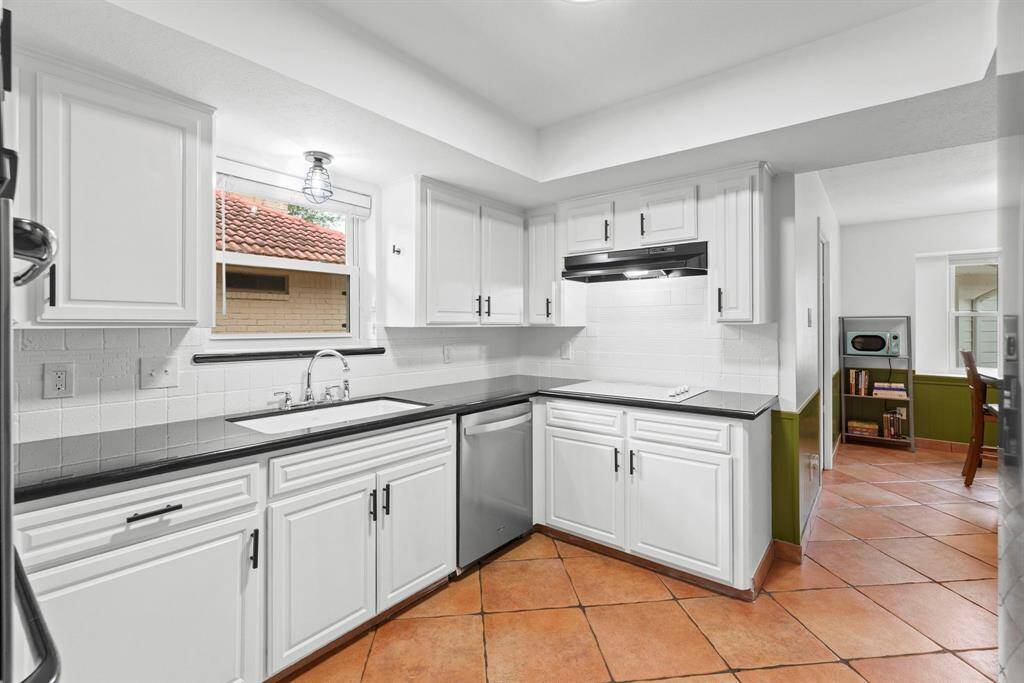 This kitchen features sleek white cabinetry, a modern stainless steel dishwasher, and granite countertops. The space is brightened by a window above the double sink and has terracotta floor tiles for a warm, inviting feel. An adjacent breakfast area is visible, enhancing the open layout.