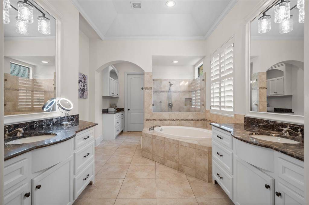 The primary bathroom features dual sinks with elegant marble countertops, a large soaking tub, a walk-in shower, and ample storage with sleek white cabinetry. Natural light streams in through stylish plantation shutters, creating a bright and inviting atmosphere.