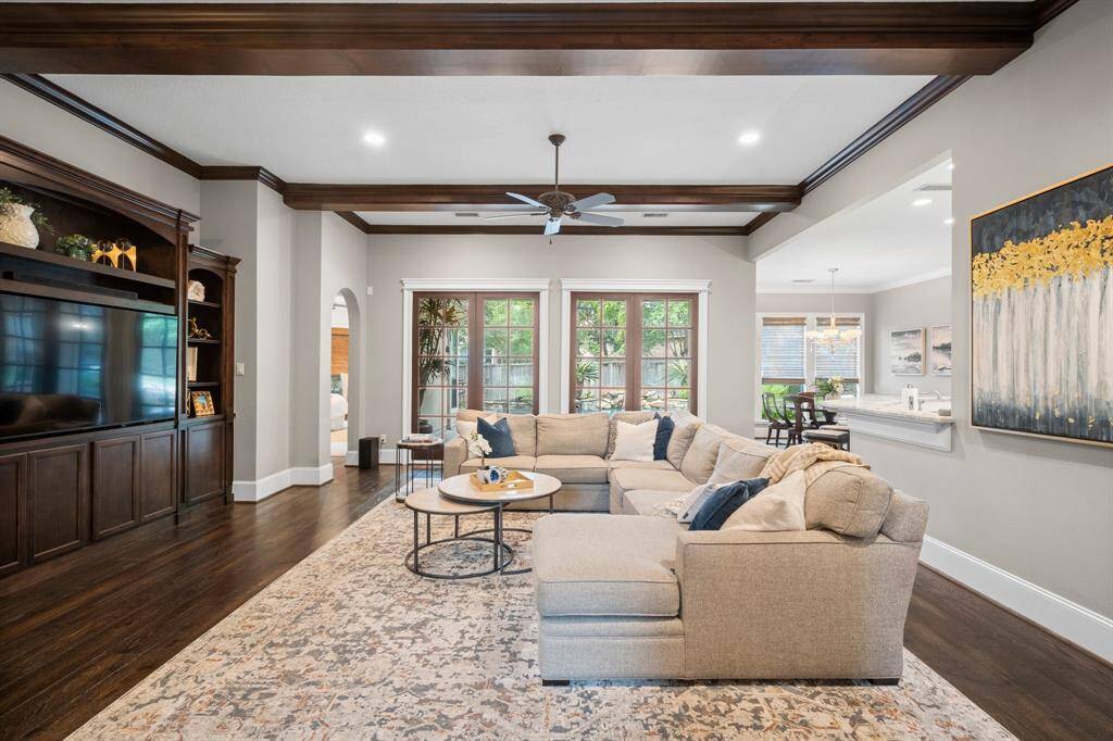 This inviting living room features elegant coffered ceiling and large windows for natural light. The open layout connects to the breakfast area and kitchen, with stylish wood flooring and a built-in entertainment center adding warmth and functionality.