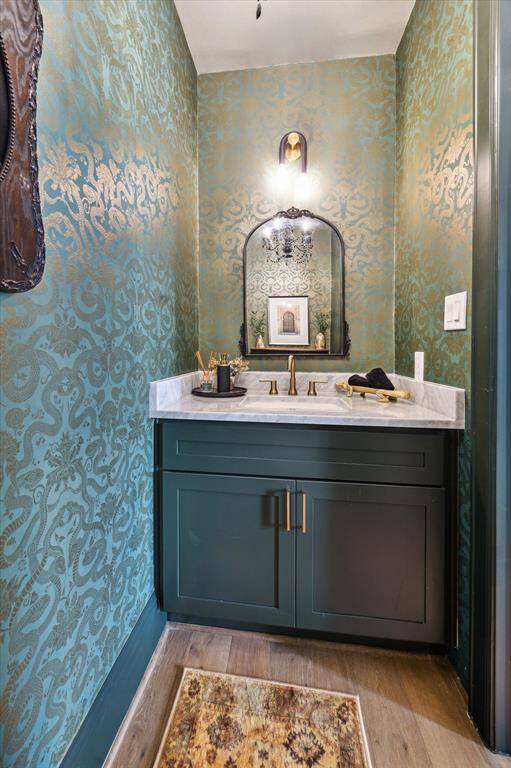 This elegant powder room features a stylish vanity with a beautiful countertop and brass fixtures. The walls are adorned with a sophisticated, patterned wallpaper in green and gold tones. A classic mirror and warm lighting add a touch of luxury, while the flooring complements the overall design.