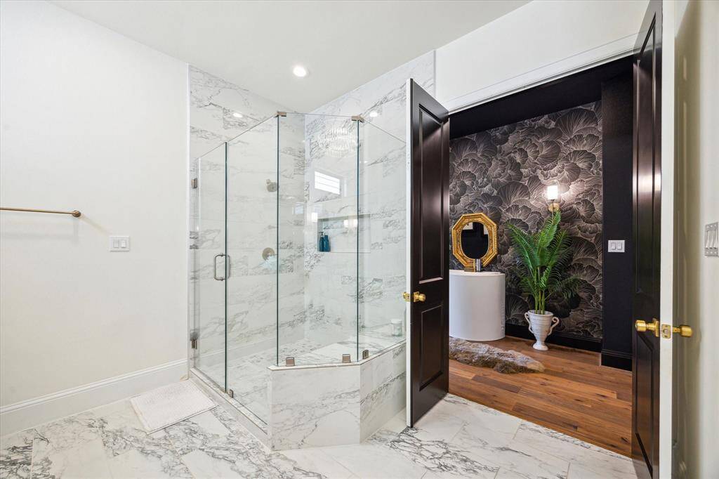 This elegant bathroom features a spacious, glass-enclosed shower with marble walls and flooring. It opens to a chic dressing area with dark floral wallpaper, a stylish mirror, and a warm wood floor, creating a luxurious, modern feel.