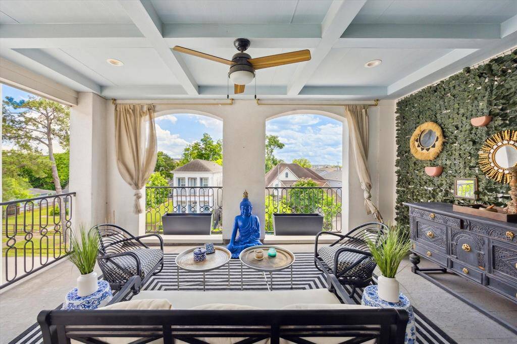 Covered outdoor terrace with a ceiling fan, stylish seating, and a serene atmosphere. It boasts a decorative wall, lush greenery, and a peaceful view, ideal for relaxation or entertaining.