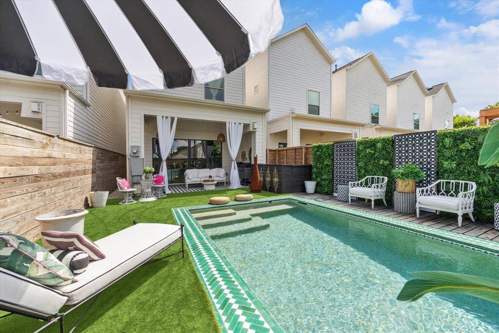 Chic outdoor space featuring a stylish pool with a morrocan tile border, lush artificial grass, and modern lounge seating. The area includes a covered patio with curtains, perfect for relaxation or entertaining, surrounded by privacy fencing and greenery.
