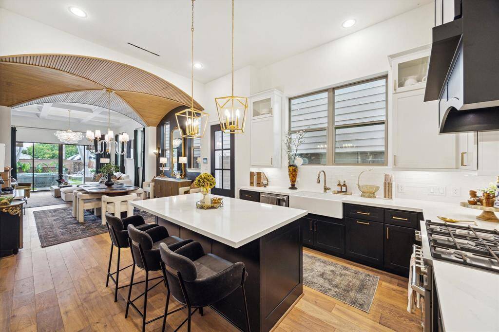 Open-concept kitchen and dining area. The kitchen features a large island with seating, sleek black and white cabinetry, and elegant light fixtures. The dining space flows seamlessly into a cozy living area with large windows offering plenty of natural light. Wood flooring and tasteful decor add warmth and sophistication.