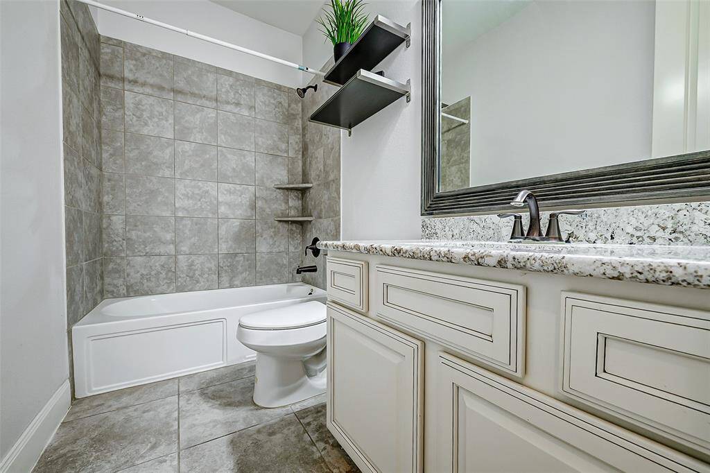 This secondary bathroom features a modern design with a tiled bathtub-shower combo, elegant granite countertop, stylish cabinetry, and a large framed mirror, creating a sleek and functional space. It is located at the front of the home next to the front bedroom.