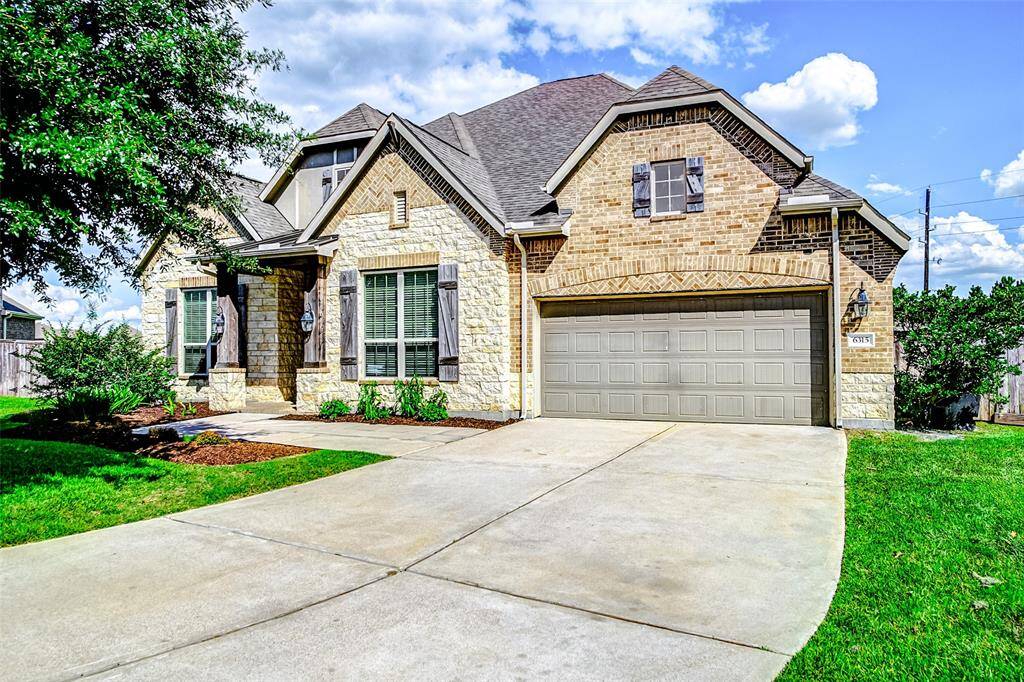 This home features a mix of brick and stone exterior, with a spacious driveway leading to a two-car garage. The house has a classic architectural design with a gabled roof and large windows, offering plenty of natural light throughout the home.