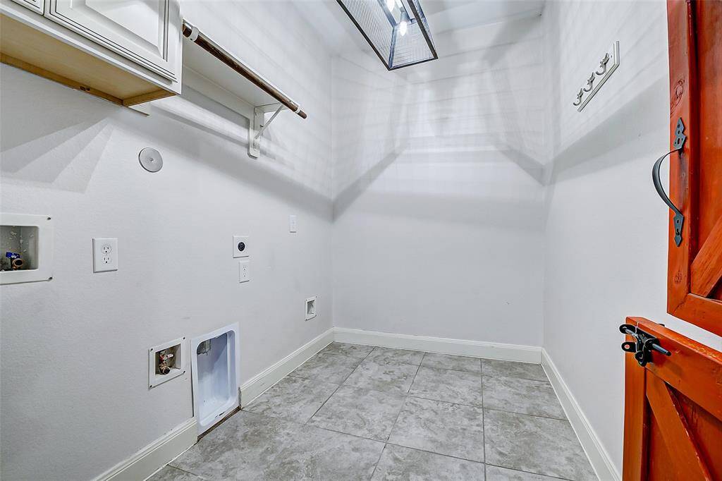 The is a spacious laundry room with tile flooring, hookups for a washer and dryer, a wooden shelf, and a small cabinet for storage. There's ample light and a convenient hanging rod.