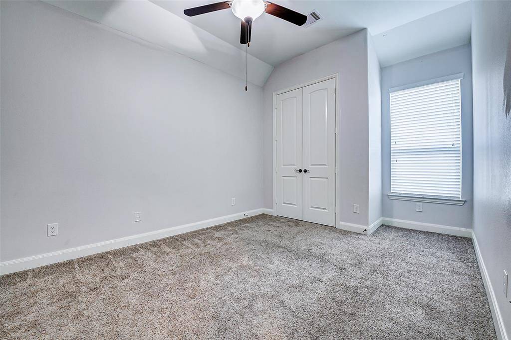 This room features soft gray walls and plush carpet, offering a cozy atmosphere. It has a ceiling fan, a double-door closet, and a window with blinds, providing ample natural light. Perfect for a bedroom or home office.