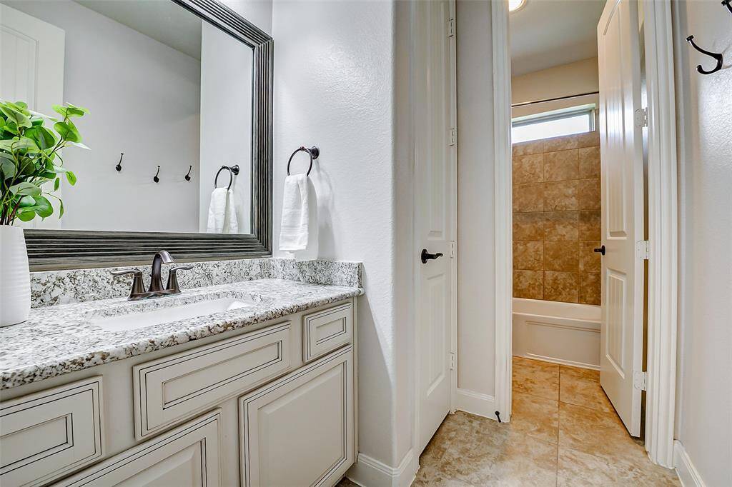 This is a modern bathroom featuring a granite countertop with a single sink, elegant cabinetry, and a large framed mirror. The space includes a tiled bathtub area, neutral tones, and stylish fixtures for a clean, contemporary look.