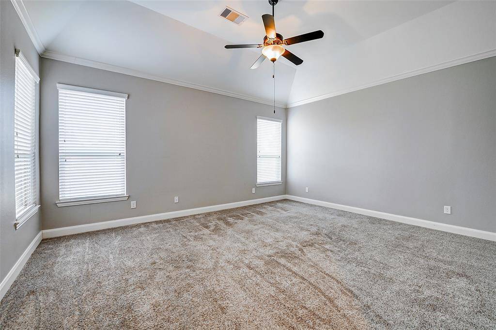 Spacious primary bedroom suite with neutral gray walls and plush carpeting. It features a tray ceiling with a modern ceiling fan, and two large windows that provide natural light.