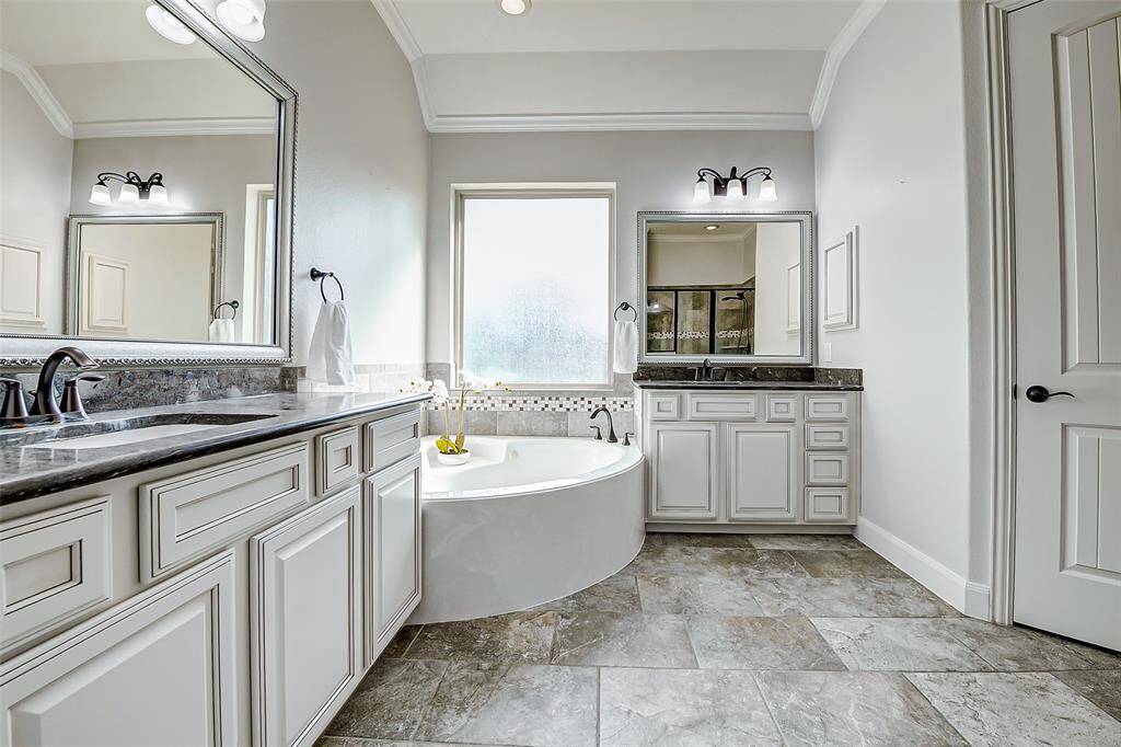 The primary bathroom features a spacious layout with elegant white cabinetry, dual sinks with dark countertops, and a luxurious soaking tub. The large window allows natural light to brighten the room, and the neutral tile flooring complements the classic design.