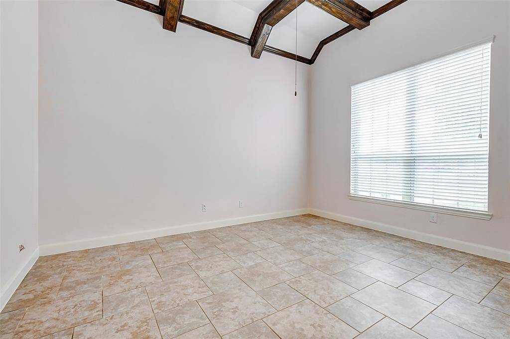 Bright home office with high ceilings featuring exposed wooden beams. Large window with blinds allows natural light to fill the space. The room has neutral-toned tile flooring, creating a clean and open atmosphere.