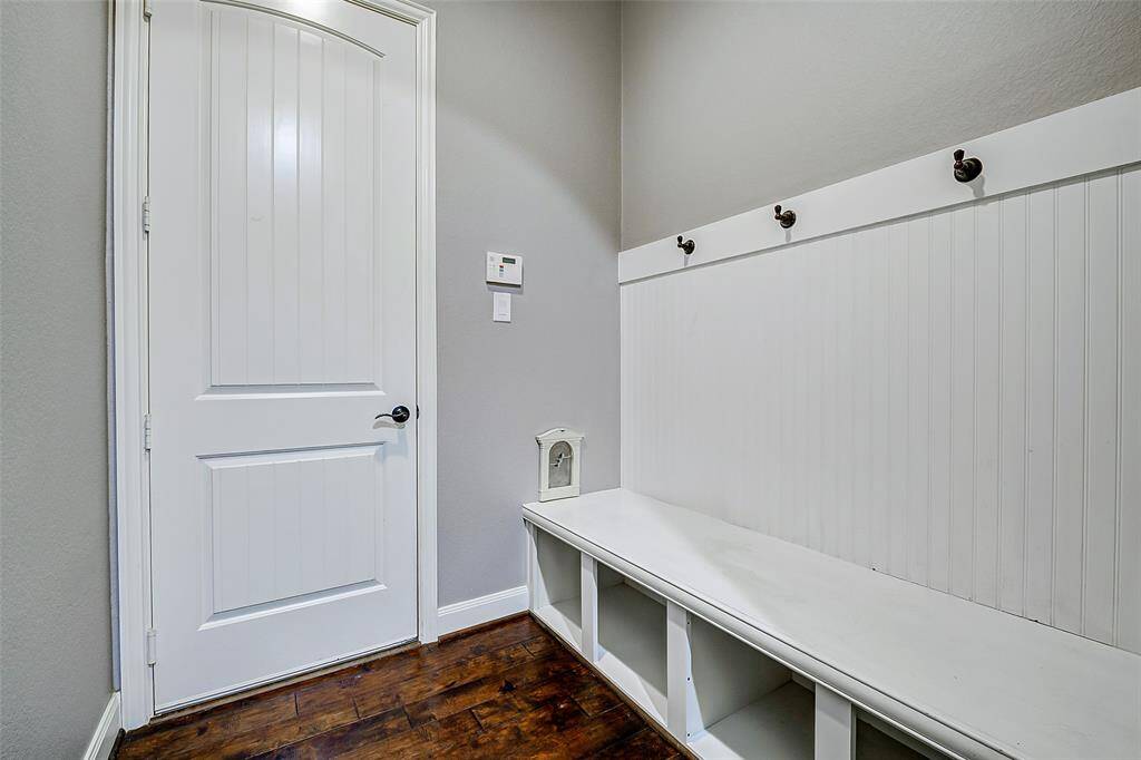 This photo features a mudroom with a built-in bench and storage cubbies beneath. The wall has hooks for hanging coats, and the door opens to the garage, all set against light gray walls and dark wood flooring.