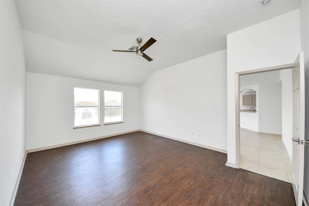 This room features a spacious layout with dark hardwood floors, two large windows for natural light, a ceiling fan, and an adjoining doorway leading to a tiled area, possibly a kitchen or hallway.