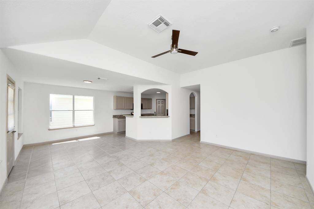 This photo shows a spacious, open-concept living area with tiled floors and neutral walls, featuring a ceiling fan and large windows for natural light. The room flows into a kitchen area with a breakfast bar and modern cabinetry.