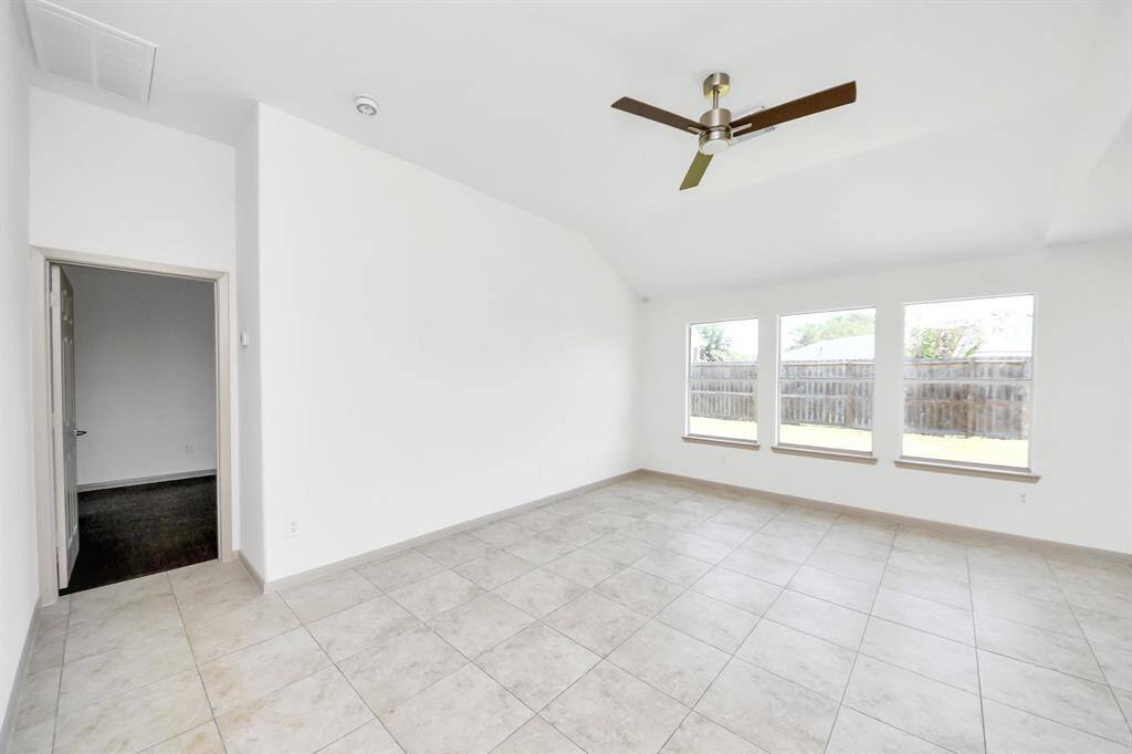 This spacious room features high ceilings, large windows offering plenty of natural light, tile flooring, and a ceiling fan. The neutral palette provides a versatile backdrop, and the view of the fenced backyard adds a touch of privacy.
