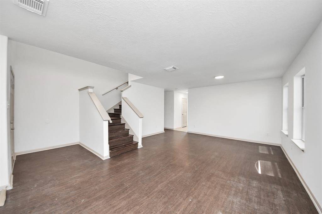 This photo shows a spacious living area with dark hardwood floors and neutral walls. The room features a staircase with a modern railing, multiple windows for natural light, and a door leading to another area of the house.