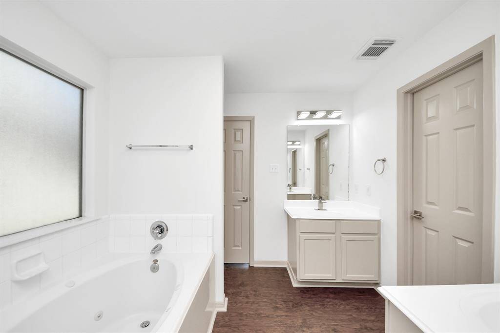 This bathroom features a large soaking tub beneath a frosted window, a spacious vanity with ample storage, and a sleek mirror with modern lighting. The space is bright and airy, with neutral tones and wood flooring.