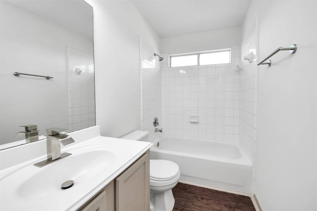 This bathroom features a clean, modern design with white cabinetry and walls, a large wall mirror, and a bathtub with overhead shower. It has a sleek, integrated sink and faucet, complemented by a wood-look floor.