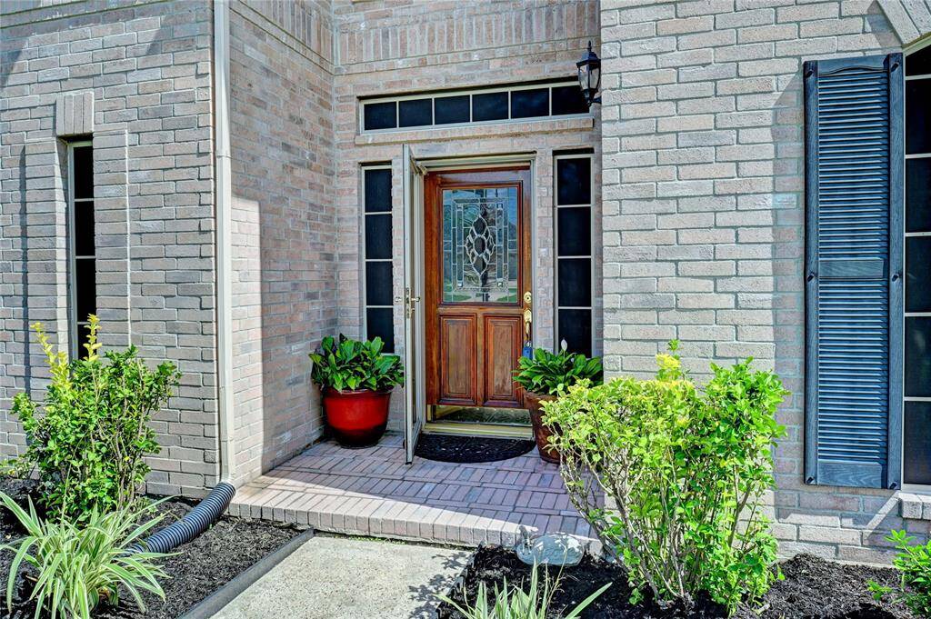 This welcoming entryway features a classic wooden door with decorative glass, framed by tall windows. The brick exterior is complemented by vibrant potted plants and manicured shrubs, creating an inviting first impression.
