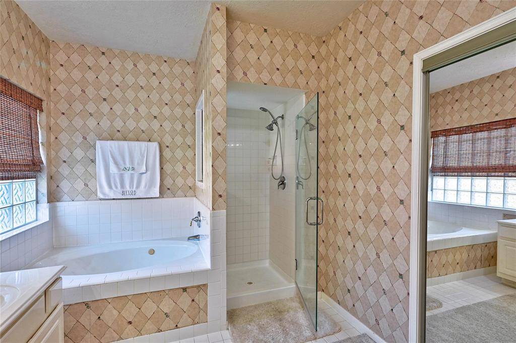 This bathroom features a soaking tub, a separate glass-enclosed shower, and a dual-sink vanity. It has patterned tile walls and large windows with natural light. Large walk in closet.