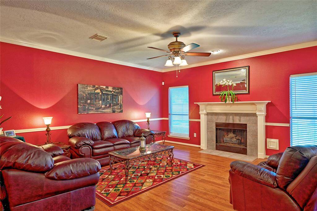 This inviting living room features rich red walls and warm solid flooring, complemented by a cozy fireplace.  A ceiling fan and ample lighting complete the room's appeal. Open view to kitchen and breakfast area.