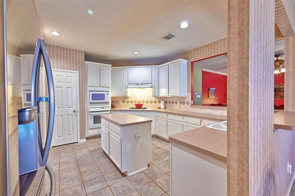 This kitchen features a bright, open layout with white cabinetry and ample counter space. It includes built-in appliances, an island, and a view into an adjoining room with a vibrant color scheme. The tiled floor and recessed lighting add a modern touch.