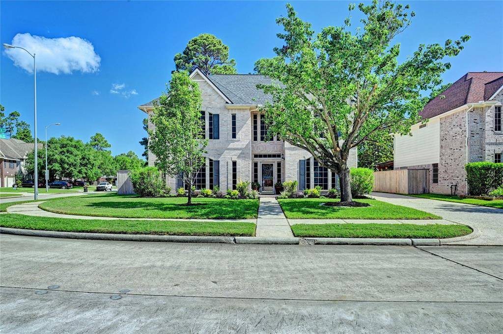 This photo shows a charming two-story brick house on a corner lot with a well-maintained lawn and mature trees. The home features a welcoming front entrance and is located in a Villages of Northgate Crossing