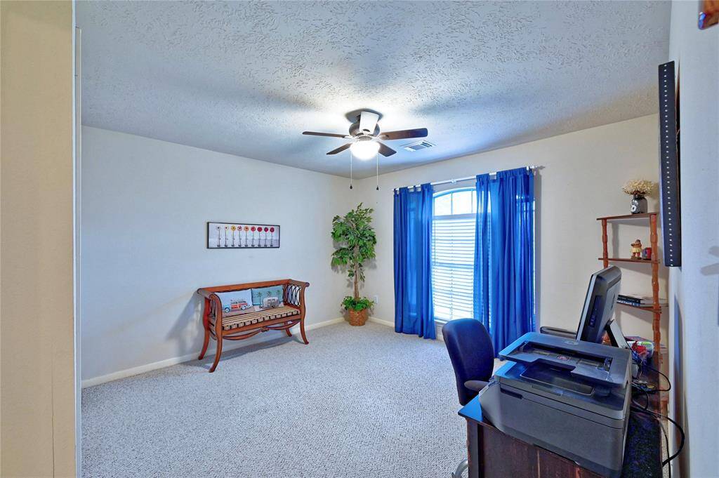 This room is a bright, spacious home office with a ceiling fan, large window with blue curtains, and comfortable carpeted flooring and walk in closet.
