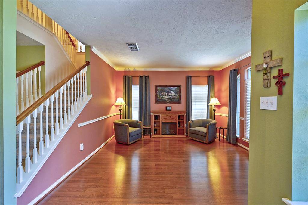 Bright and inviting formal den with warm solid flooring, a cozy seating arrangement, and ample natural light. The room features an elegant staircase and a small entertainment center, perfect for relaxation and gatherings.