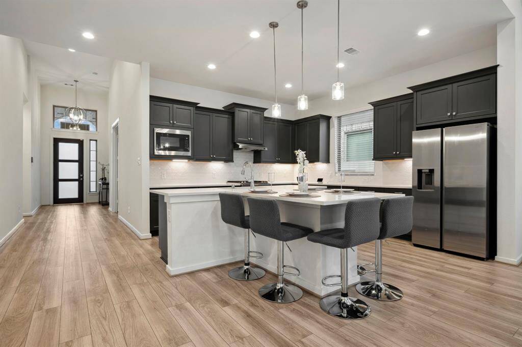 This modern kitchen features sleek black cabinetry, stainless steel appliances, and a spacious island with seating. The open layout and wood flooring create a welcoming and stylish atmosphere.