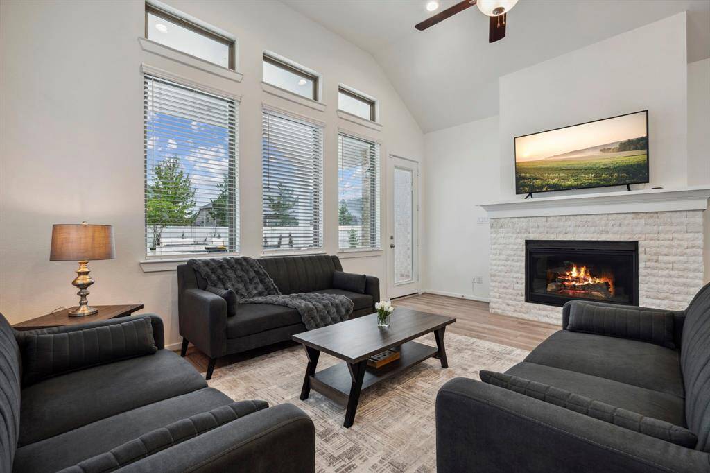 This cozy living room features a high ceiling with a ceiling fan, a modern fireplace, and a TV above the mantel. Natural light pours in through large windows, illuminating the comfortable seating area with plush sofas and a coffee table, creating an inviting atmosphere.