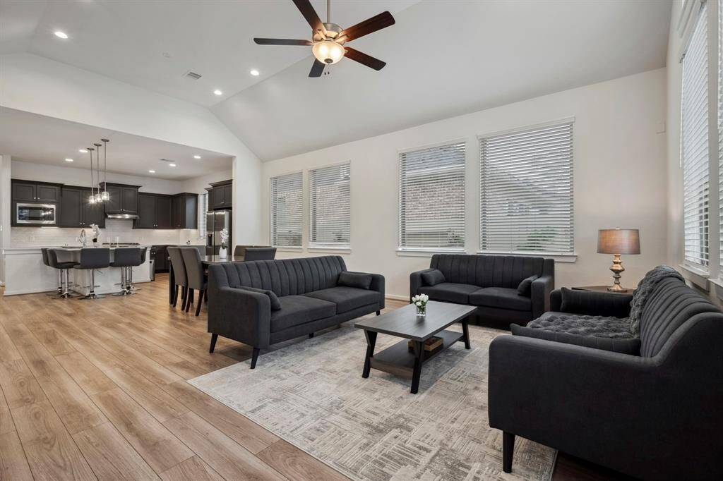 This inviting open-concept living space features a modern kitchen with dark cabinetry and a breakfast bar, a cozy dining area, and a spacious living room with ample natural light, sleek furniture, and a ceiling fan, all accented by wood-look flooring.