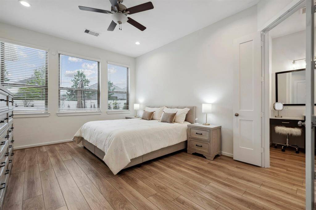 Bright and inviting bedroom with large windows, wood flooring, and a ceiling fan. Includes a cozy double bed, nightstands, and an adjacent vanity area.